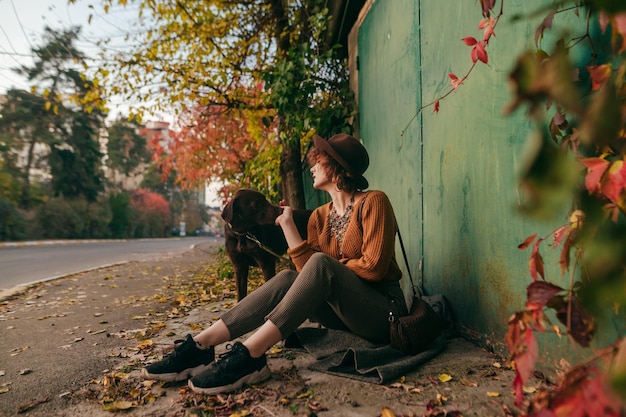 Stylish lady with dog sits on street in village with dog on leash on background of autumn landscape