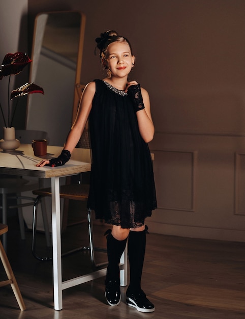 A stylish little girl in a black dress stands in the interior near the table.