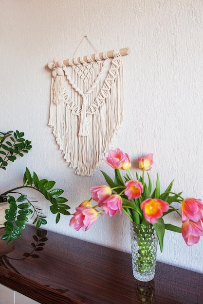 Stylish macrame wall hanging on the white wall above the chest of drawers Cozy interior of living room with flowers in vase macrame and hanging green plant Minimalistic concept of home decor