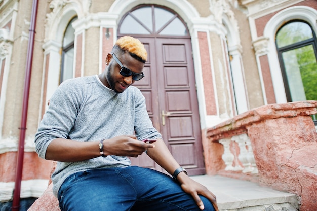 Stylish man on gray sweater and black sunglasses posed on street Fashionable black guy looking on mobile phone.