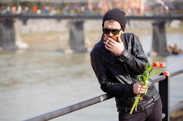 Stylish man with a beard in sunglasses with a mustache in a warm hat and leather jacket holds a bouquet of red tulips and smokes a cigarette