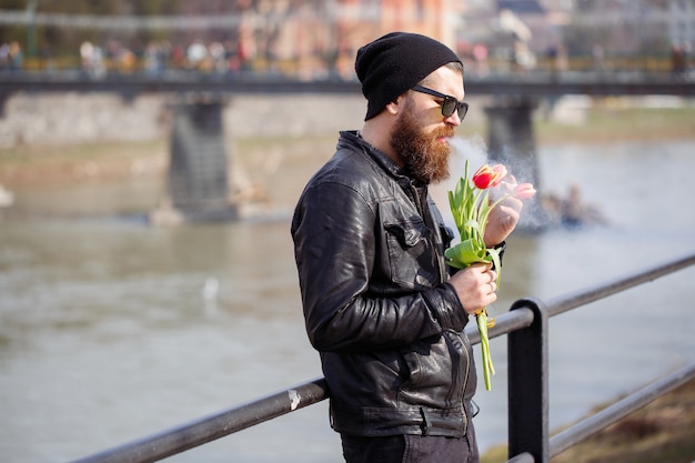 Stylish man with a beard in sunglasses with a mustache in a warm hat and leather jacket holds a bouquet of red tulips and smokes a cigarette