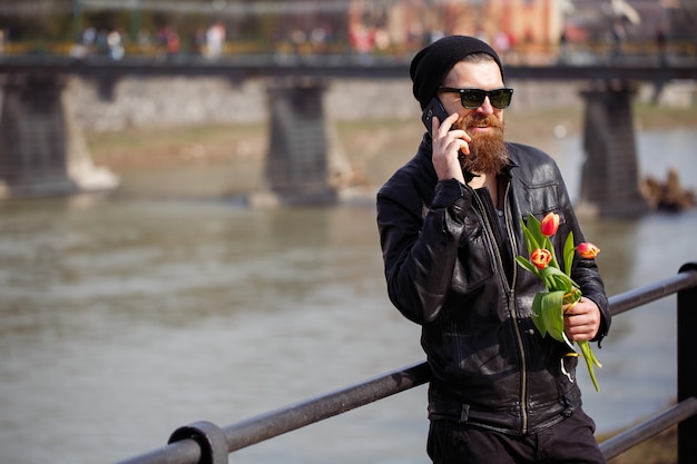 Stylish man with a long beard and mustache in a black warm hat and leather jacket holding a bouquet of red tulips and smiling speaks on the phone