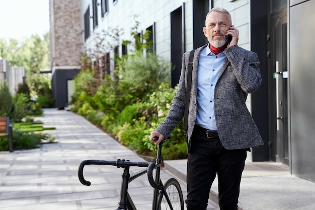 Stylish mature businessman looking away while talking on the phone standing with bicycle outdoors