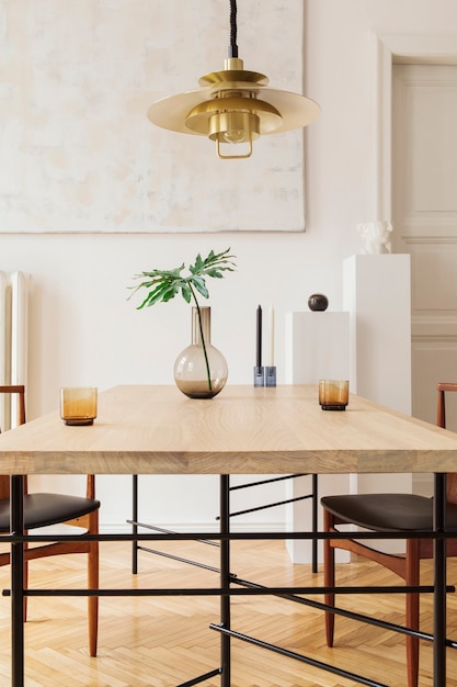 Stylish and modern dining room interior with design sharing table, chairs, gold pendant lamp, abstract paintings and elegant accessories. Tropical leafs in vase. Eclectic home decor.