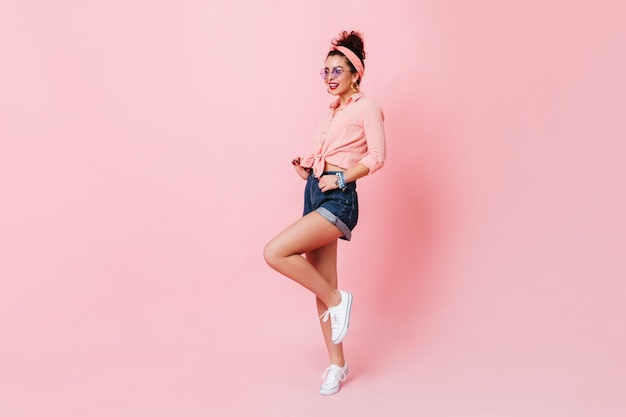 Stylish pinup girl with red lips lifts her leg on pink background Fulllength shot of woman in pink shirt and denim shorts