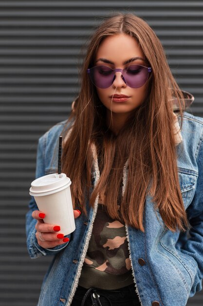 Stylish portrait hipster pretty young woman in fashionable purple glasses in a trendy blue denim jacket with a hot drink in hands near gray wall on the street. Attractive girl model resting outdoors.