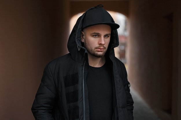 Stylish portrait of a young handsome man in a fashionable black baseball cap in a warm black hood with a hood on the street