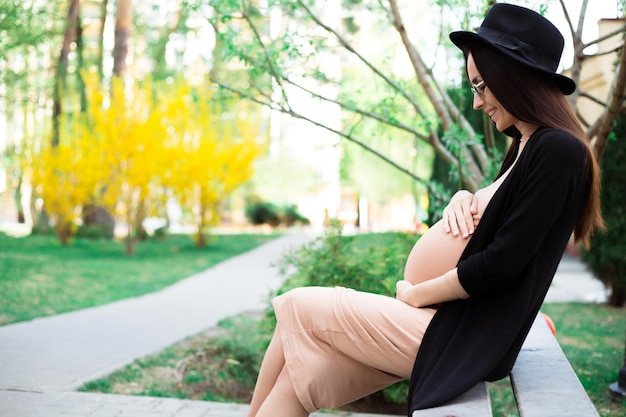 Stylish Pregnant in Hat and Sunglasses Sitting on a Bench in the City Park and Relax