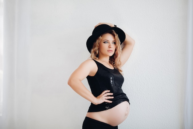 Stylish pregnant woman in black hat and sparkling top embracing her belly.