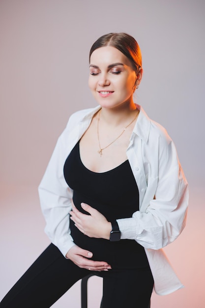 A stylish pregnant young woman in a white shirt and black pants sits on a chair and hugs her belly on a white background A pretty woman with professional makeup and casual clothes