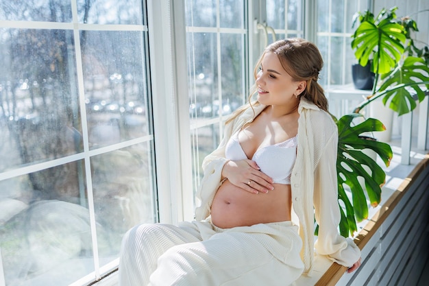 Stylish pregnant young woman in a white suit near the window and touching her belly with a gentle smile Awaiting birth Care and motherhood Love and tenderness