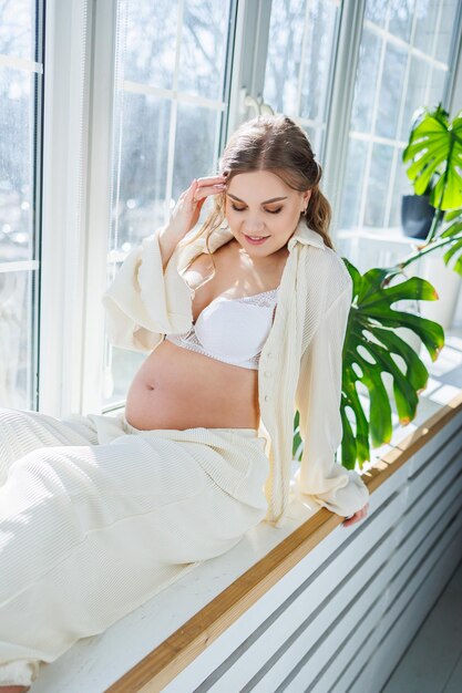 Stylish pregnant young woman in a white suit near the window and touching her belly with a gentle smile Awaiting birth Care and motherhood Love and tenderness