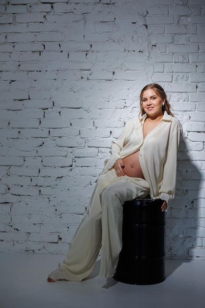 A stylish pregnant young woman in a white suit sits against a white brick wall and touches her belly with a gentle smile Awaiting birth Care and motherhood Love and tenderness