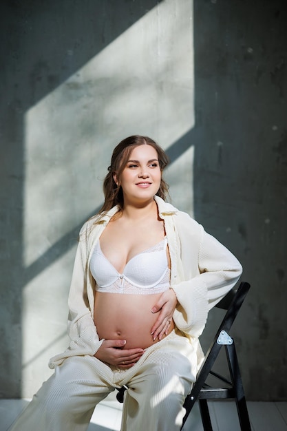 A stylish pregnant young woman in a white suit sits on a chair and touches her belly with a gentle smile Awaiting birth Care and motherhood Love and tenderness