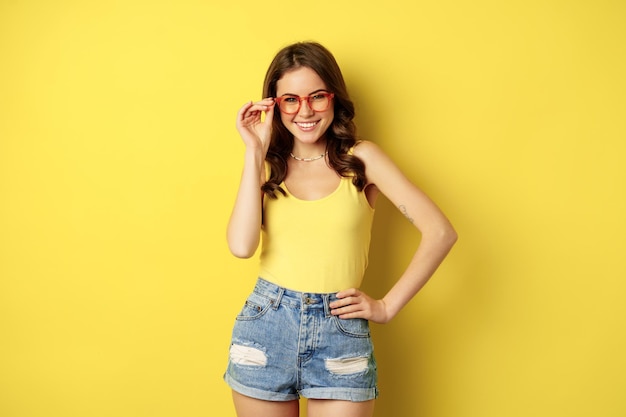 Stylish pretty woman posing in sunglasses and summer clothes, looking happy and carefree, concept of vacation and sunny days, standing over yellow background