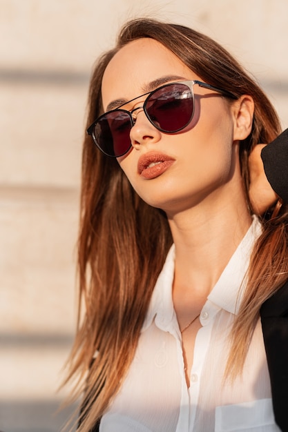 Stylish pretty woman in sunglasses in fashionable clothes on the street near a gray wall at sunset