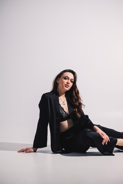 A stylish pretty young woman in a black classic suit is sitting on a white background A beautiful woman in a jacket and makeup on her face