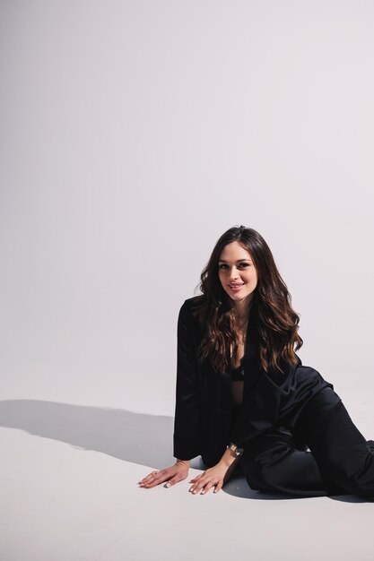 A stylish pretty young woman in a black classic suit is sitting on a white background A beautiful woman in a jacket and makeup on her face