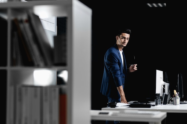 Stylish professional architect in glasses dressed in a blue checkered jacket stands next to the desk with computer in the office .