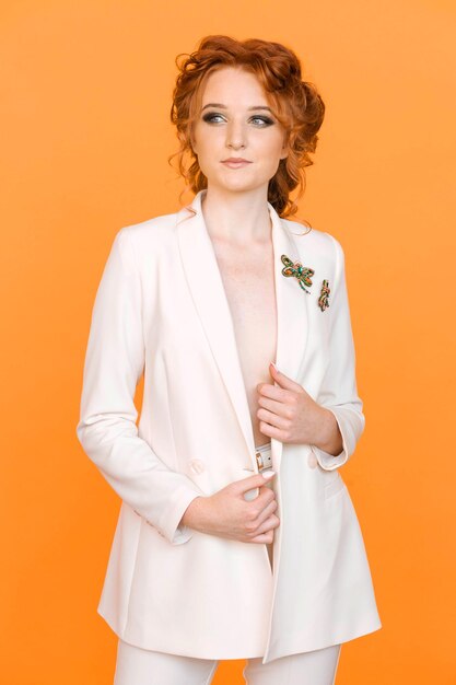 Stylish redhaired girl posing on an orange background in a white jacket with a brooch in the shape of a dragonfly