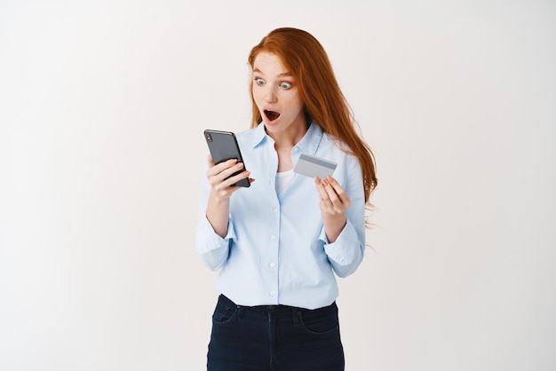 Stylish redhead girl staring surprised at smartphone screen while shopping online holding plastic card reaction on internet special deal promotion