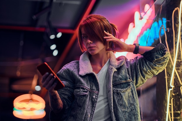 Stylish redhead girl wearing a denim coat holding a phone and looking at camera while leaning on the illuminated signboard in the night on the street.