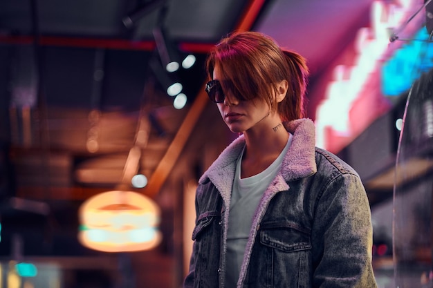 Stylish redhead girl with tattoo on her face wearing a denim coat and sunglasses standing in the night on the street. Shop signboards, neon, lights.