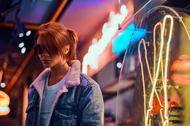 Stylish redhead girl with tattoo on her face wearing a denim coat and sunglasses standing in the night on the street. Shop signboards, neon, lights.