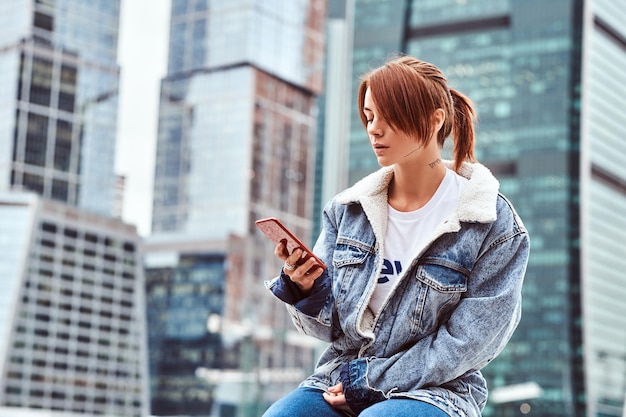 Stylish redhead hipster girl with tattoo on her face wearing denim jacket using a smartphone sitting in front of skyscrapers in Moscow city at cloudy morning.