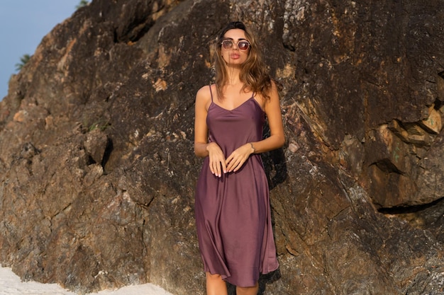 Stylish romantic tender sensual woman in silk dress and sunglasses on the beach at sunset