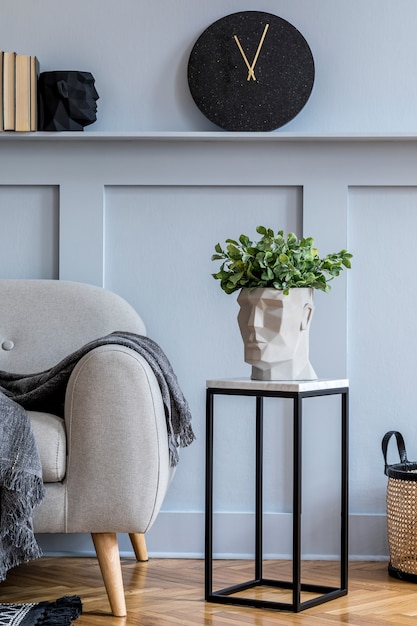 Stylish scandinavian interior of living room with grey sofa, plaid, black clock, wood paneling with shelf, marble stool, plants and elegant personal accessories in design home decor.