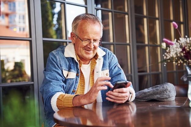 Stylish senior in fashionable clothes and in glasses sits in the cafe with phone.