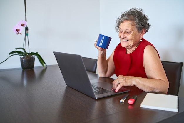 Stylish senior woman using laptop during a video call with her family and holding a blue mug