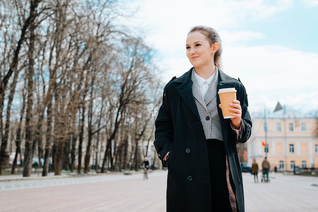 Stylish smiling young woman in trendy black coat standing in park and looking away Positive pretty woman with cup of coffee outdoors copy space