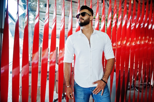 Stylish tall man in white shirt, jeans and sunglasses posed at street of city Beard attractive guy against red wall.