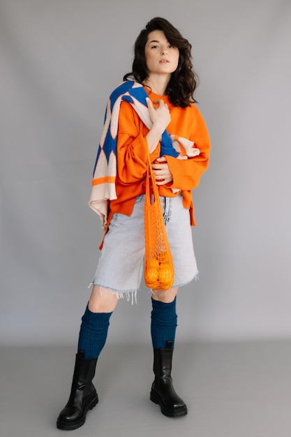 Stylish woman dressed in orange knitted jacket posing for a photo in a photo studio on a gray paper background Bright emotions poses modern concept