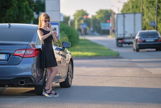 Stylish woman driver standing near her vehicle talking on cellphone on city street in summer