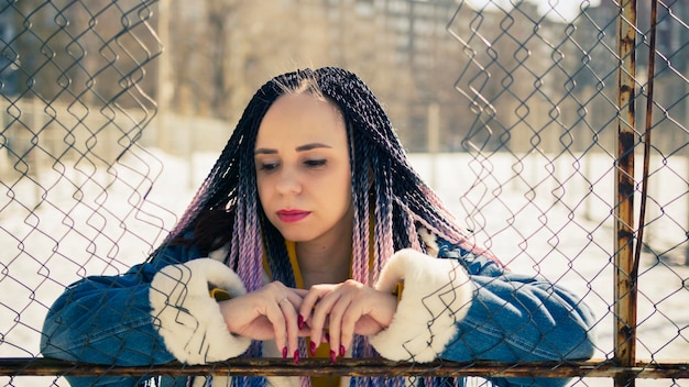 Stylish woman leaning on metal fence Serious young female in warm clothes looking away while leaning on net fence on city street