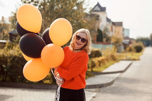 Stylish woman in orange sweater sunglasses holds orange and black balloons celebrating halloween out...