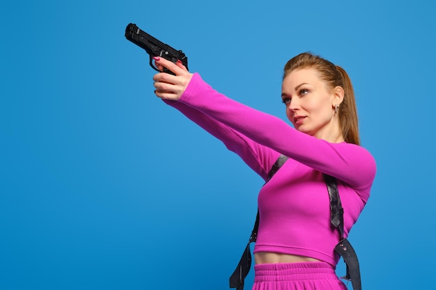 Stylish woman in purple knitted pantsuit aims a gun over blue studio background Fashion police and accurate hit on target concept