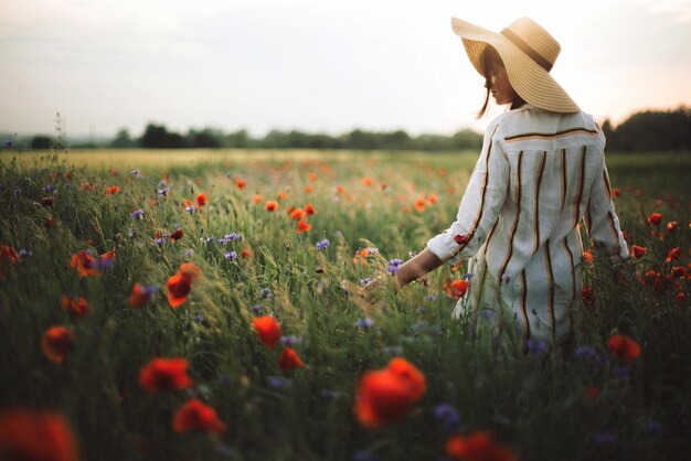 Stylish woman in rustic linen dress walking in summer meadow among poppy and wildflowers in sunset light Atmospheric authentic momentCopy space Girl in hat in countryside Rural slow life