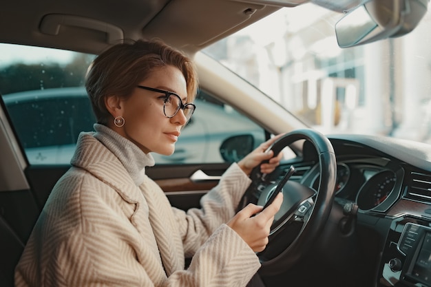 stylish woman sitting in car dressed in coat winter style and glasses using smartphone