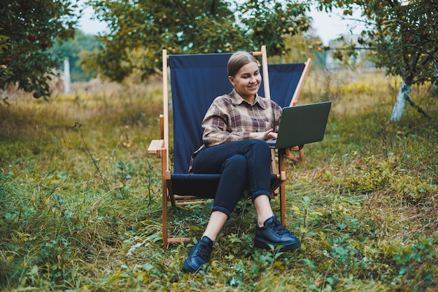 Stylish woman sitting on a chair near green plants in the garden and browsing the internet on a laptop while looking to the side Remote work female freelancer