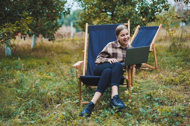 Stylish woman sitting on a chair near green plants in the garden and browsing the internet on a laptop while looking to the side Remote work female freelancer