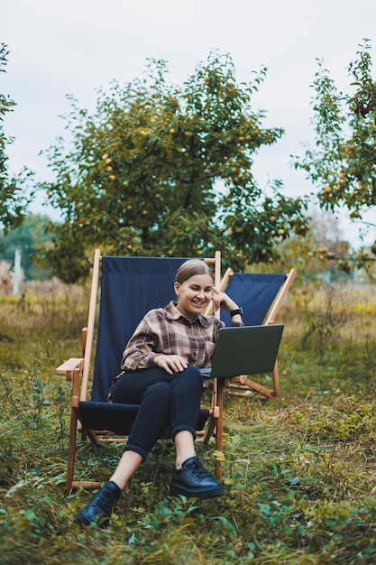 Stylish woman sitting on a chair near green plants in the garden and browsing the internet on a laptop while looking to the side Remote work female freelancer