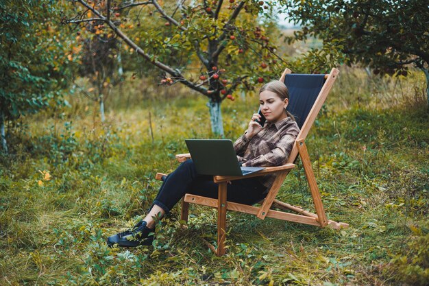 Stylish woman sitting on a chair near green plants in the garden and browsing the internet on a laptop while looking to the side Remote work female freelancer