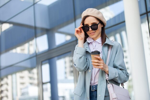 Stylish woman in sunglasses and beret holds a cup of coffee on the background of windows of the business center Fashion beauty lifestyle