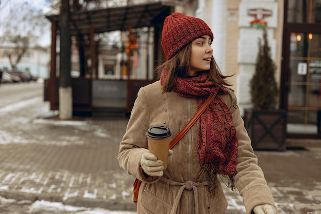 Stylish woman in warm hat and coat with cup of takeaway coffee crossing road in city