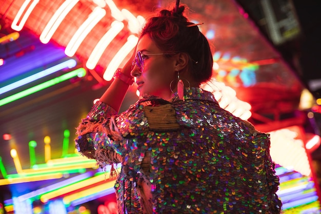 Stylish woman wearing jacket with shining sequins on the city street with neon lights